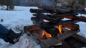 Eine Person sitzt am Lagerfeuer, im Hintergrund schneebedeckte Berge.