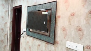 A room with patterned wallpaper, a mounted television, a door, and a power outlet. - Hotel Tulsi Palace (Jaipur)