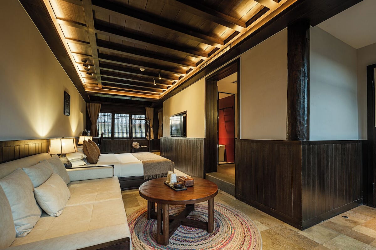 Quarto de hotel moderno com teto de madeira, um sofá, uma mesa redonda de madeira e vista para o exterior pela janela.