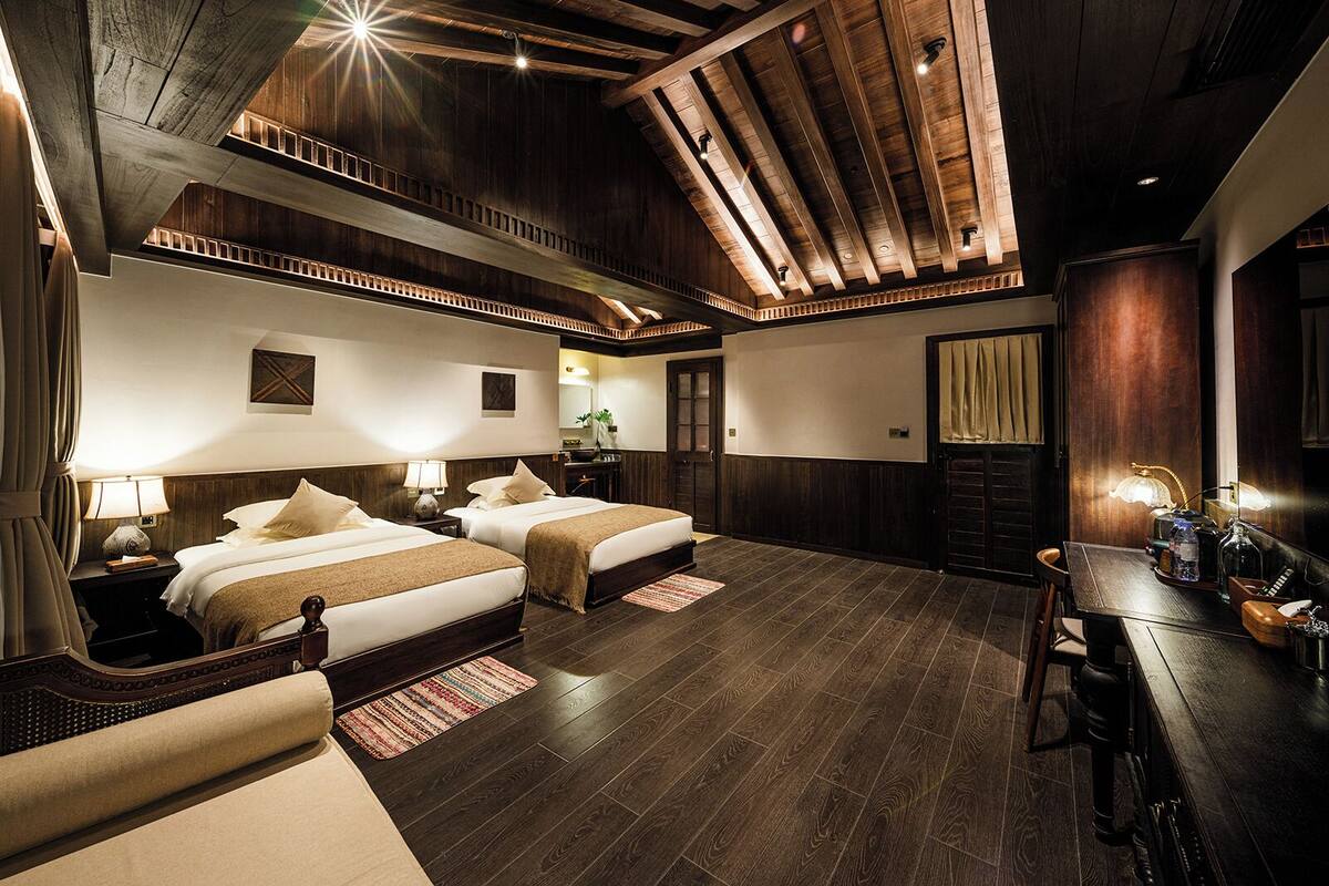 Quarto de hotel com duas camas, piso de madeira, uma mesa de jantar e teto alto com vigas aparentes.