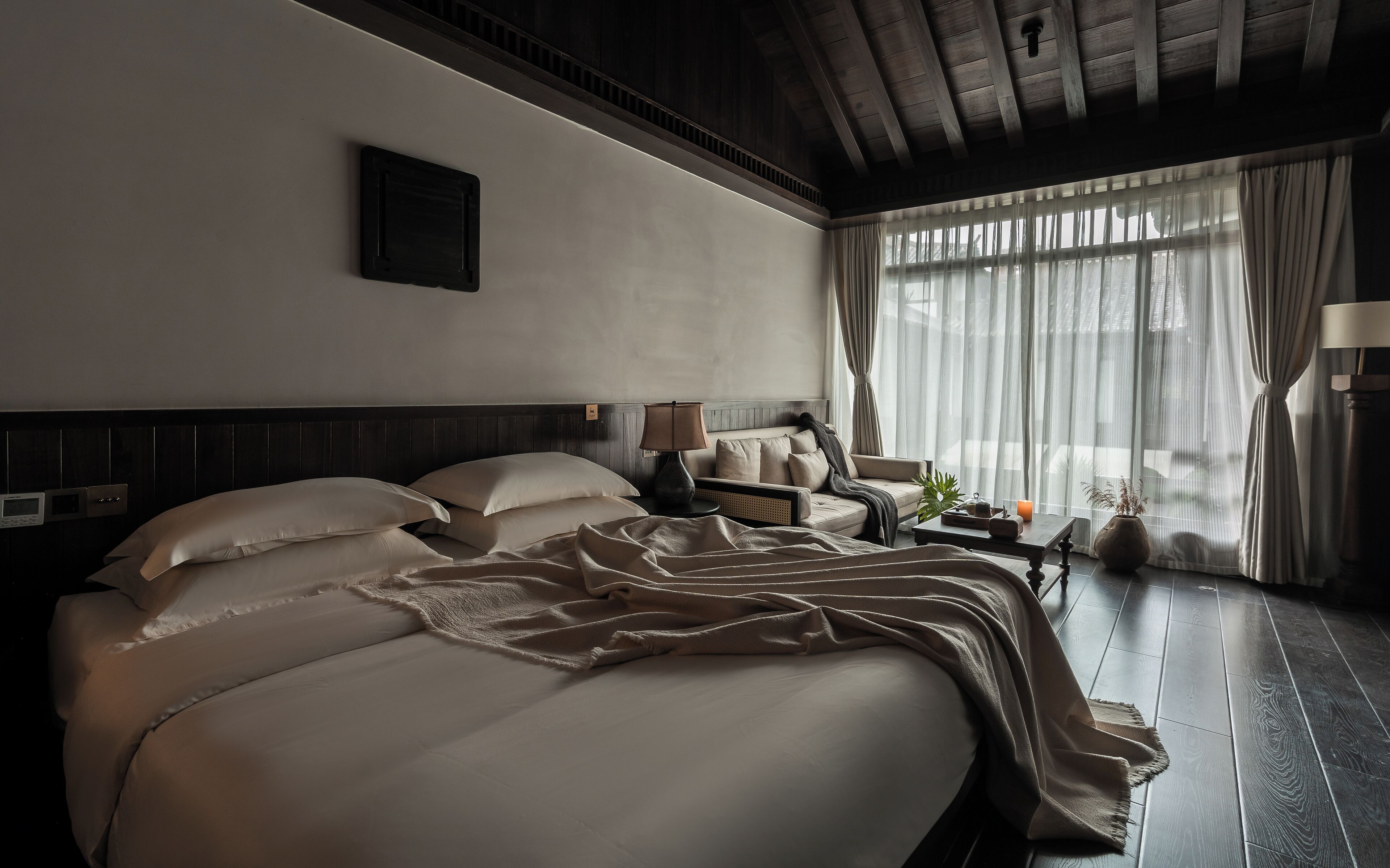 A hotel room with a large bed, a bedside table with a lamp, a small table with a candle, and a view of the outdoors through sheer curtains.