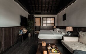 Habitación de hotel con cama, sofá, una mesa de centro de madera, planta, jarra y televisor montado en la pared.