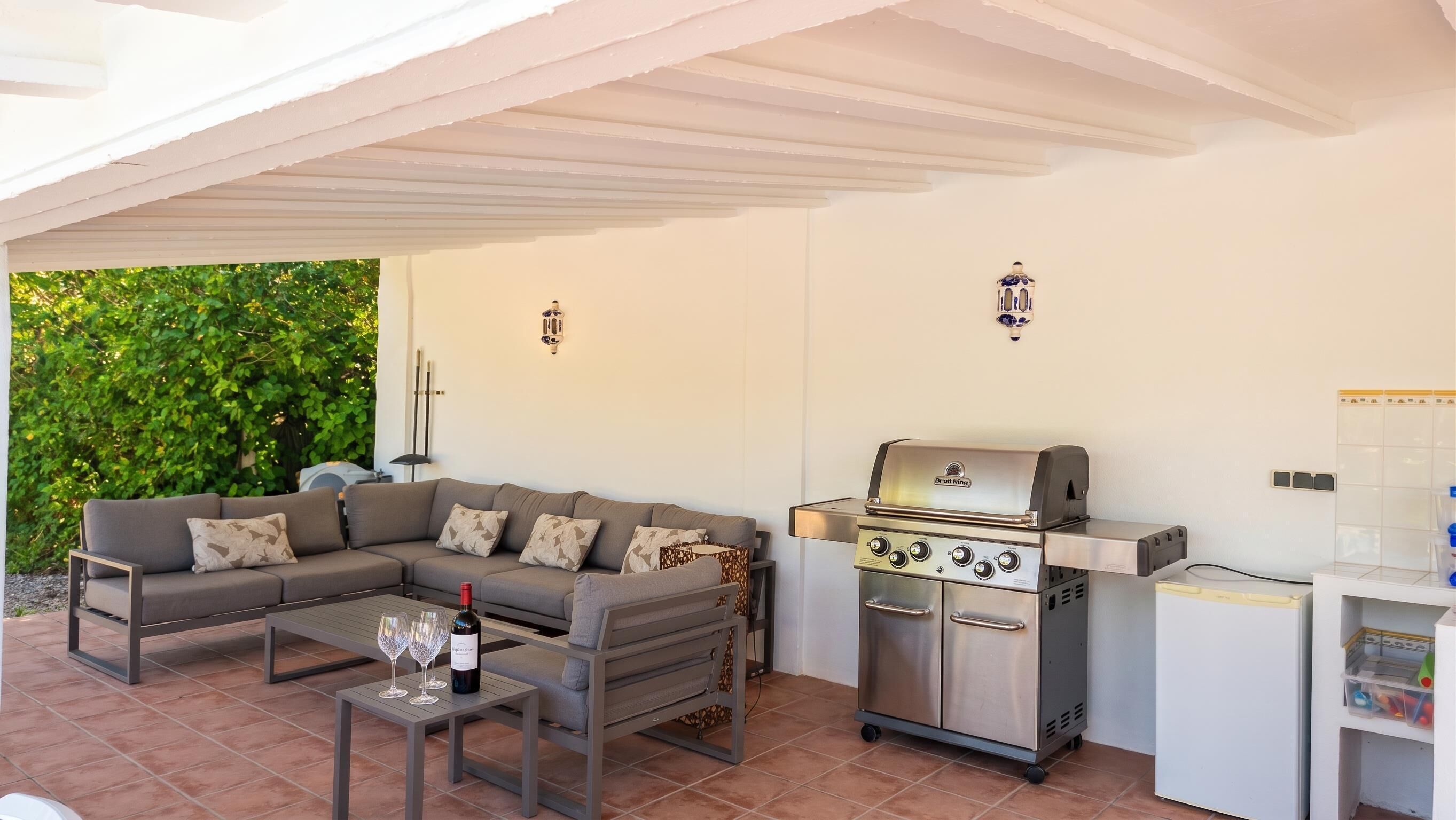 Ein überdachter Außenbereich mit einer Sitzecke, einem Sofa, einem Tisch mit Weingläsern und einem Grill.