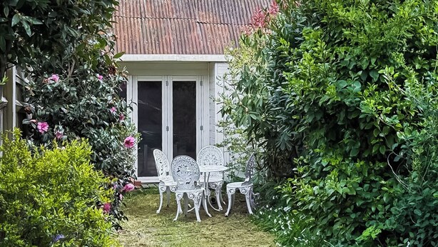 Um jardim com cerca de metal branca, um pequeno edifício branco com telhado vermelho e uma mesa com cadeiras.