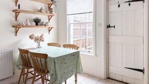 Uma sala de jantar com uma mesa posta para quatro pessoas, cadeiras de madeira, um vaso com flores secas e prateleiras acima da mesa.