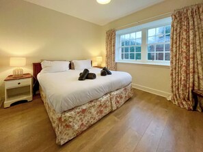 Un lit bien fait, avec du linge de lit blanc et une couvre-lit à motifs floraux, une table de chevet avec une lampe et une fenêtre avec des rideaux.