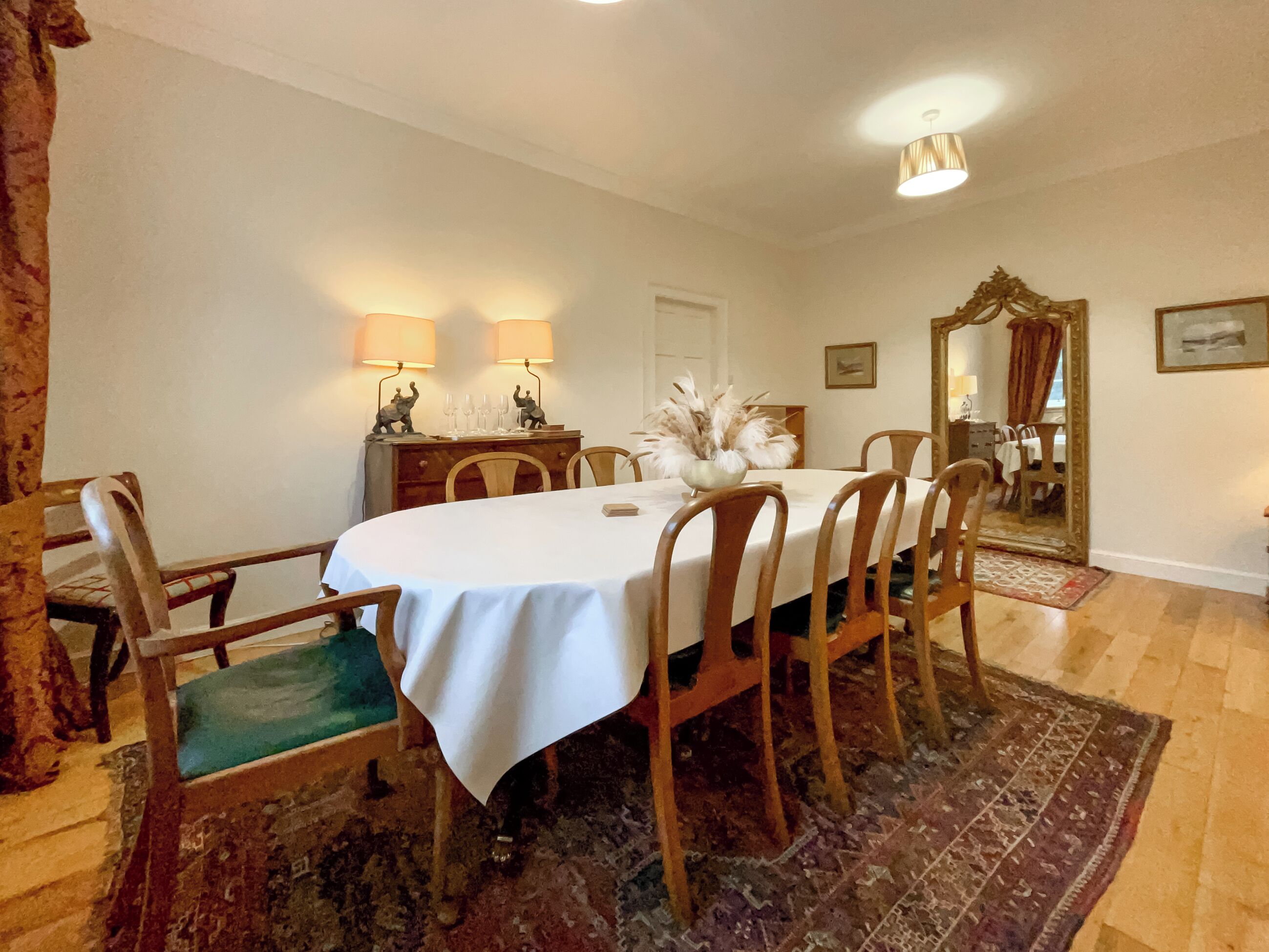 Een eetkamer met een tafel voor vier personen, houten stoelen, een spiegel en een lamp op een dressoir.