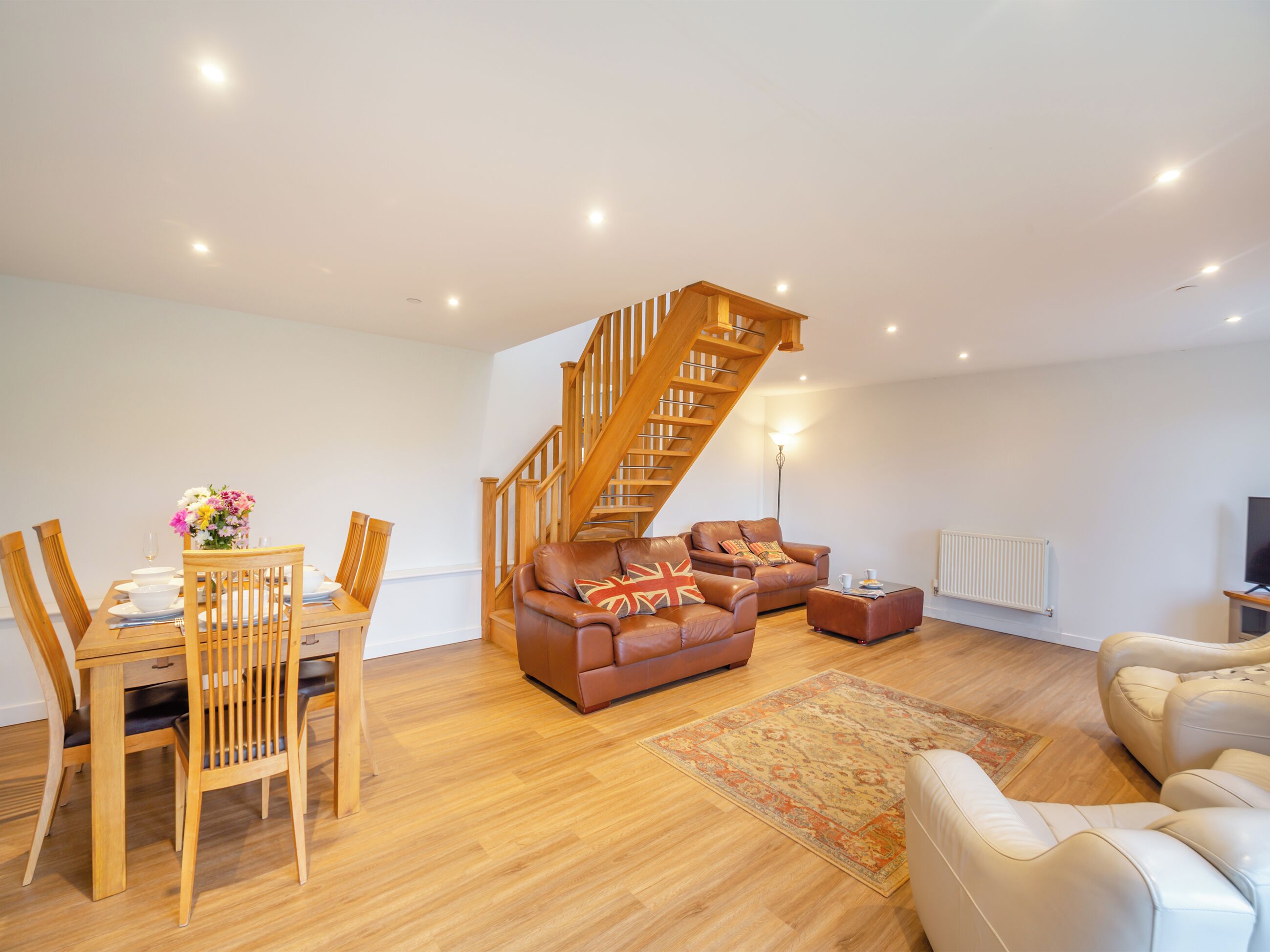 A spacious living room with a wooden staircase, a dining area with wooden chairs and a table, and a sofa with a Union Jack pillow.