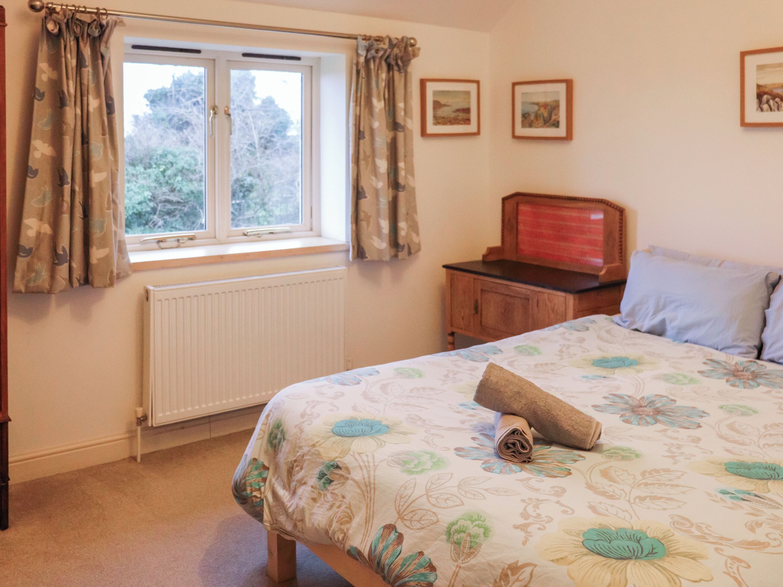 A bedroom with a bed, a window with curtains, a radiator, a wooden cabinet, and two framed pictures on the wall.
