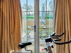 A room with a stationary bike, a door leading to an outdoor area with a table and chairs, and curtains on the windows. - 3 bedroom accommodation in Bradford Abbas (Bradford Abbas)