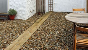 A patio area with a wooden table and chairs, a potted plant, a wooden path, and a white wall with a ladder. - Stable Cottage - UK51203 (Fishguard)