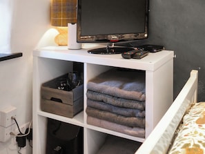 A neatly organized room with a bed, a TV on a stand, a remote control, and a shelf with folded towels and a storage box. - Maids Cottage - UK51204 (Fishguard)