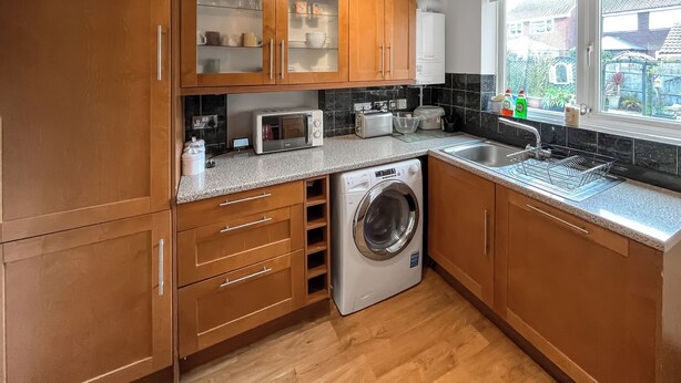 A kitchen with wooden cabinets, a microwave, a washing machine, and a window with a view of houses. - 3 bedroom accommodation in Sittingbourne (Sittingbourne)