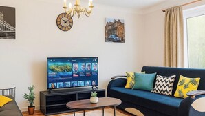 A modern living room with a flat-screen TV, a sofa, a coffee table, a chandelier, and a clock. - 3 bedroom accommodation in Sittingbourne (Sittingbourne)
