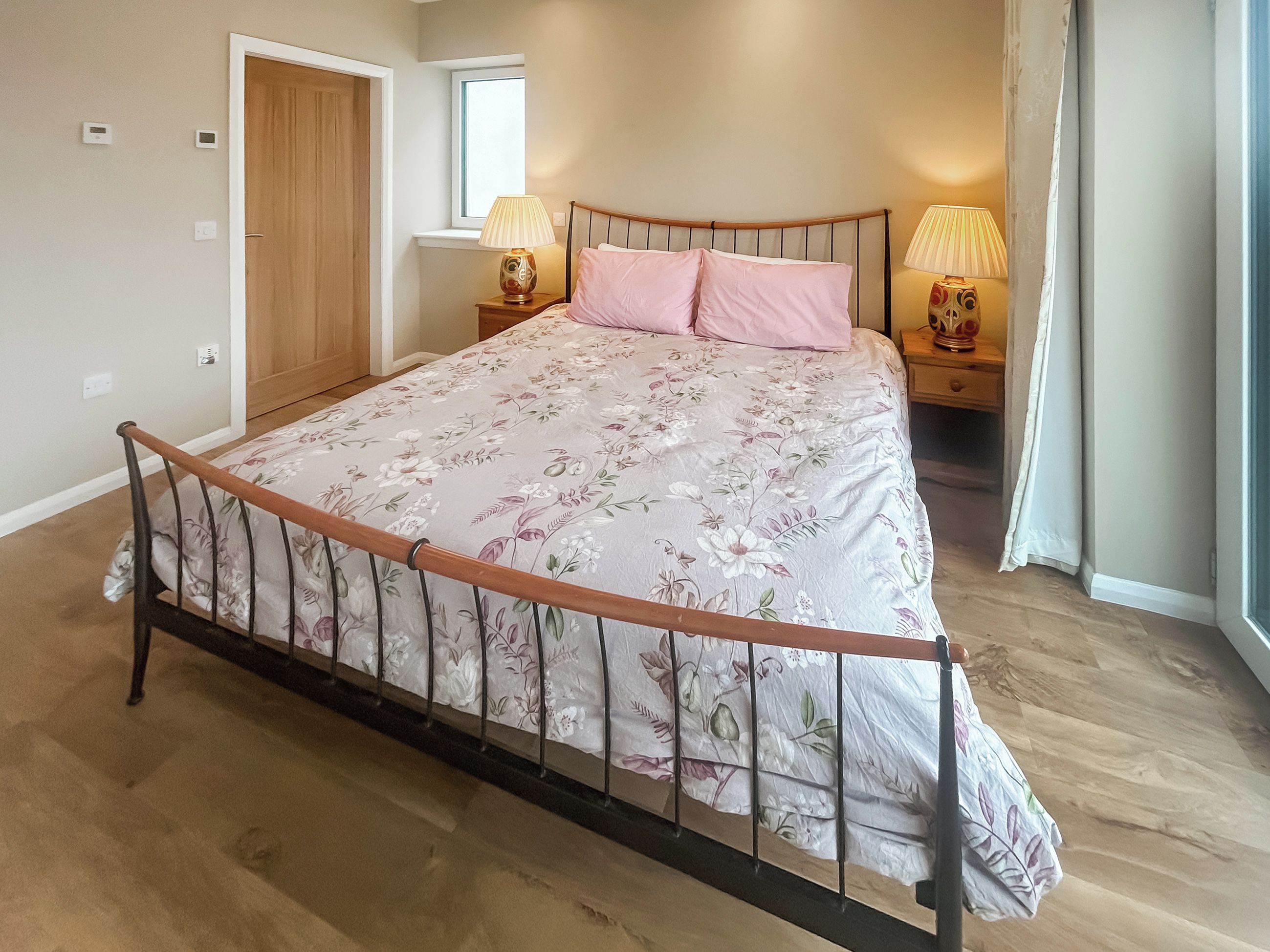 Un dormitorio con estructura de cama de madera, ropa de cama floral, una ventana y dos lámparas de noche.