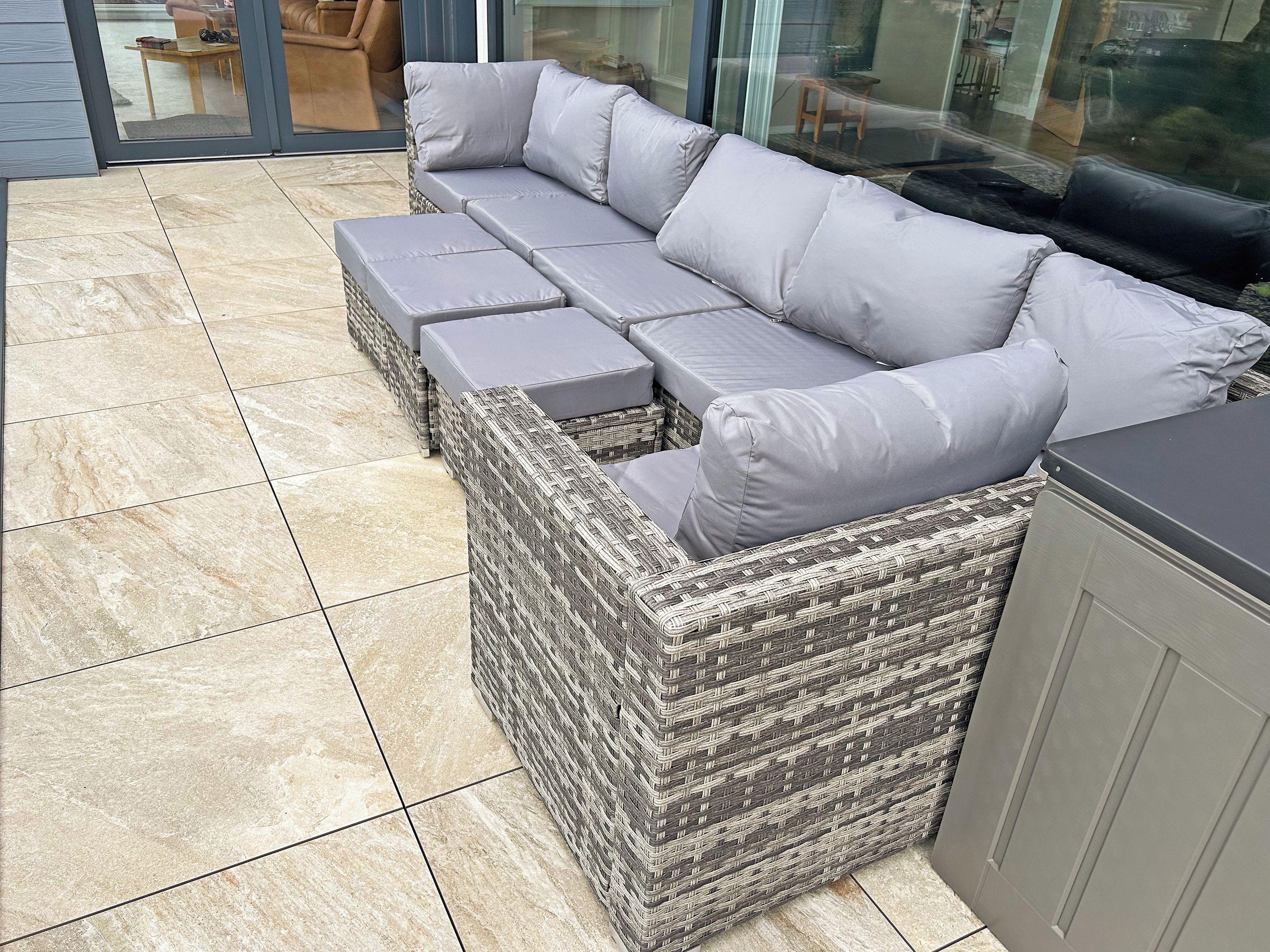A wicker sectional sofa with grey cushions, a tiled patio floor, and a glass door leading to an outdoor area.