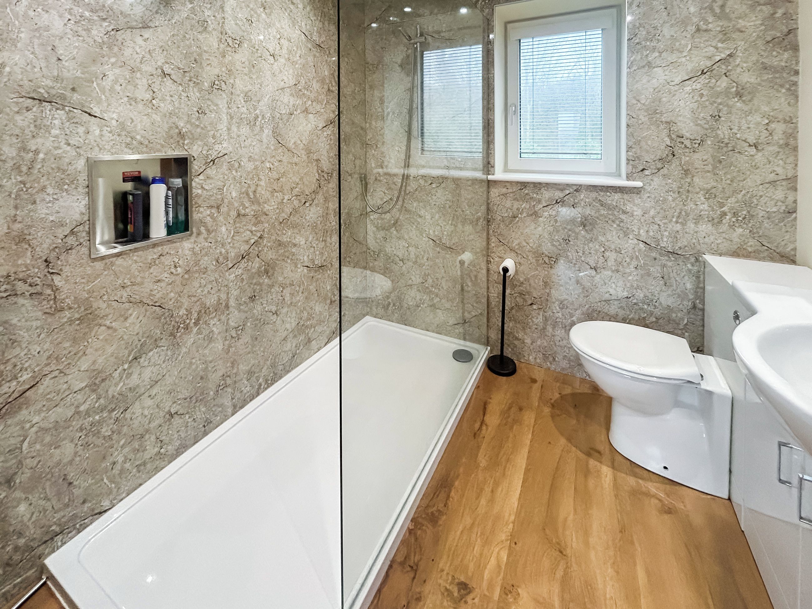 A bathroom with a marble wall, a white bathtub, a toilet, and a window with blinds.