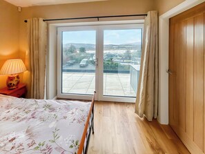 A bedroom with a bed, a nightstand, a lamp, a window with curtains, and a view of a residential area.