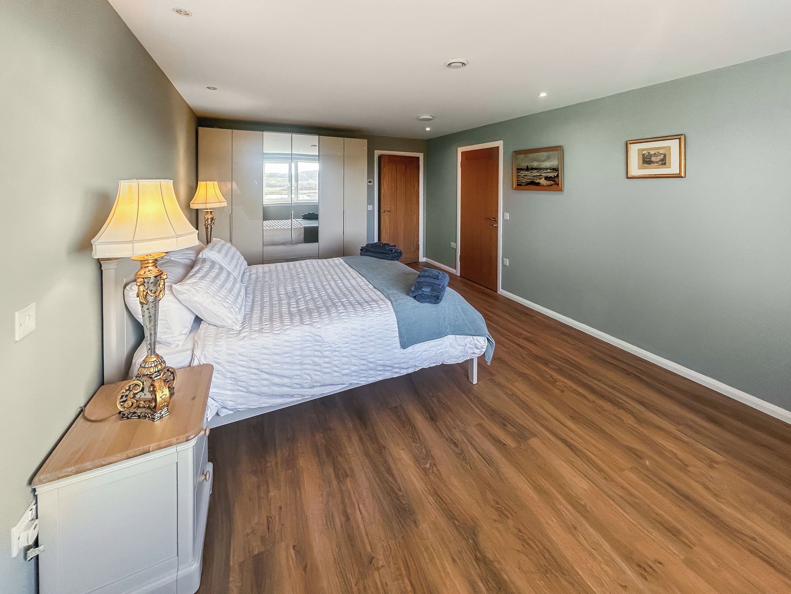 A bedroom with a bed, two bedside lamps, a wooden nightstand, a wardrobe, and a door leading to another room.