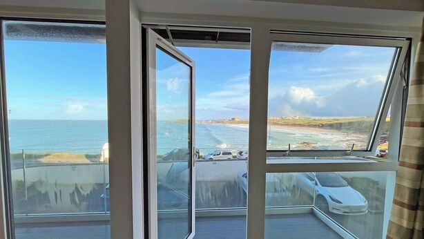 A balcony with a view of the sea and beach, a car parked on the street, and a building in the distance. - 1 bedroom accommodation in Newquay (Newquay)