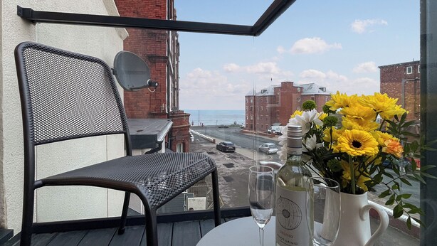 Ein Balkon mit einem Stuhl, einem Tisch mit Weinflasche und Glas sowie einer Vase mit Blumen.