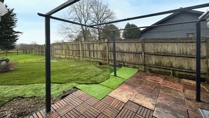 Ein Garten mit einer Holzterrasse, einer Metallpergola und einem eingezäunten Bereich.