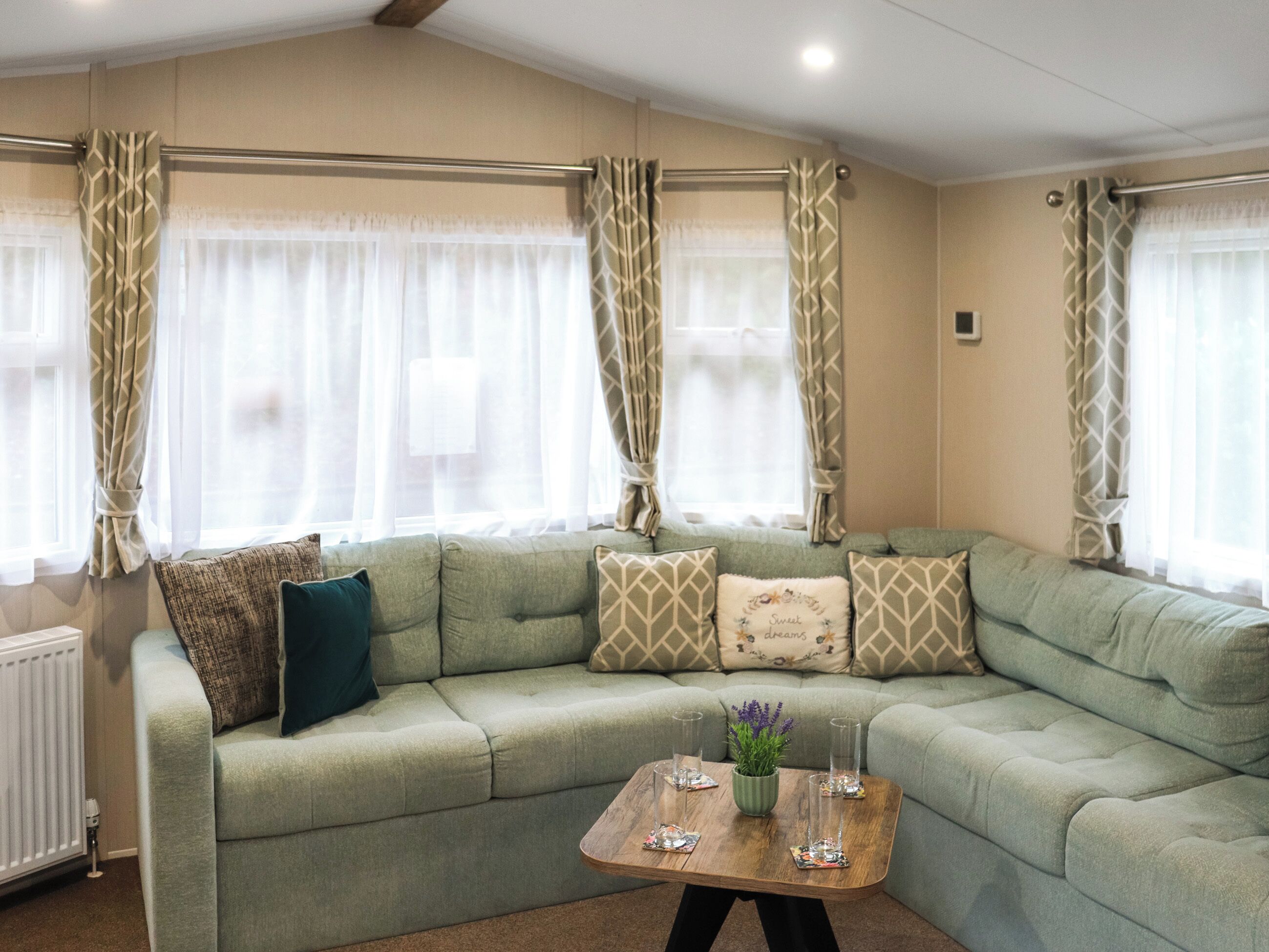 A green sofa with patterned cushions, a wooden coffee table with glassware and a plant, and sheer curtains on windows.
