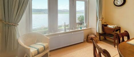 A room with a view of a body of water, a wicker chair, a wooden table, and a wall clock.