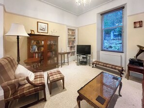 A living room with a sofa, armchair, ottoman, coffee table, television, bookshelf, and a window with blinds. - Welcome to Failte, a wonderful four-bedroom house. (Tighnabruaich)