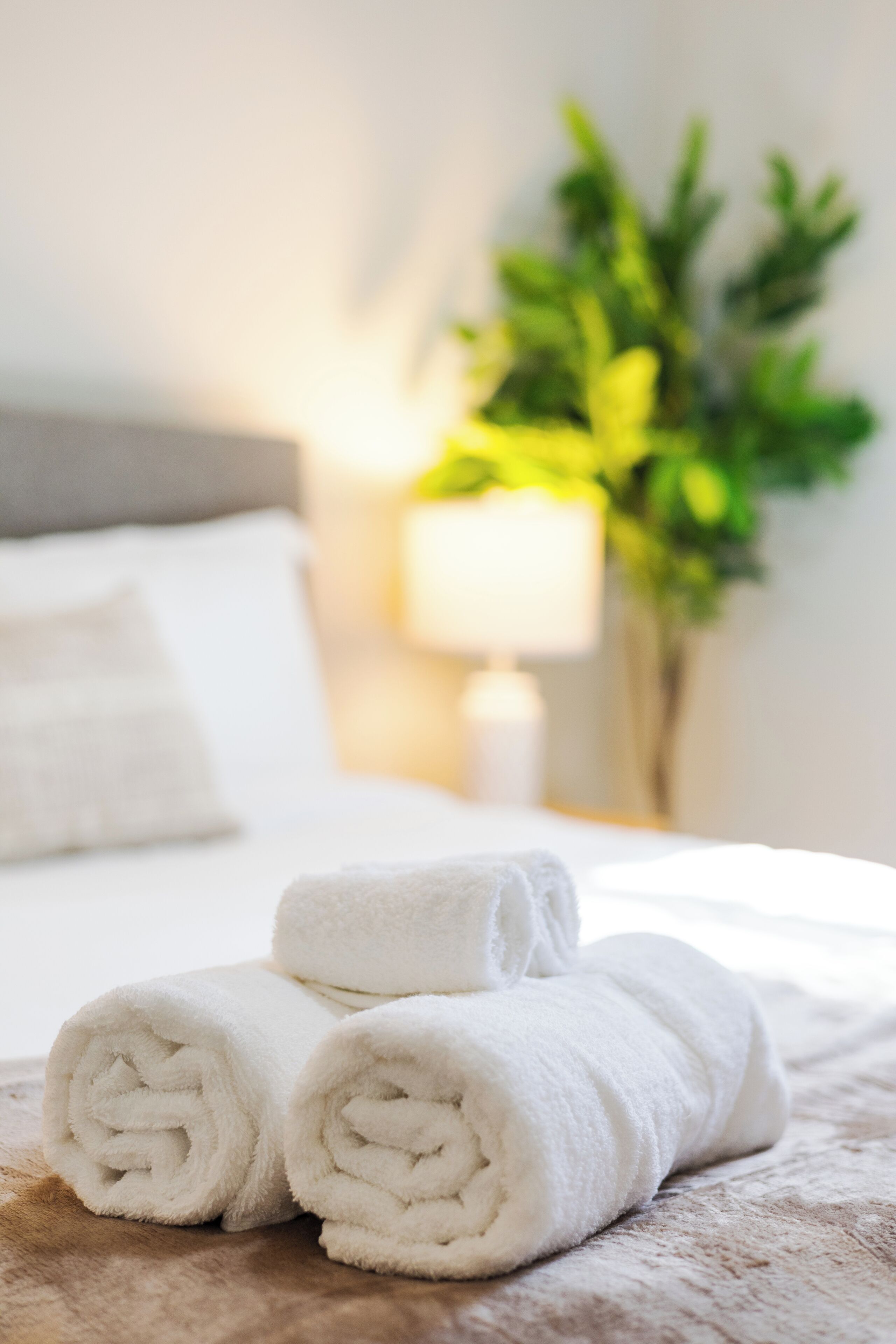 Een opgemaakt bed met opgerolde witte handdoeken en een lamp op de achtergrond.