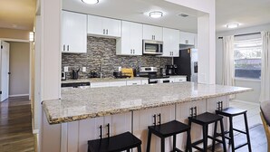 Uma cozinha moderna com armários brancos, bancada de granito e backsplash de pedra. Há três banquetas pretas e um micro-ondas.