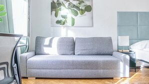 Et moderne værelse med en grå sofa, en seng med et blåt sengegærde, og et billed af eukalyptusblade på væggen.