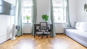 Et værelse med parketgulv i træ, en grå sofa, en sort kontorstol, et lille bord med en potteplante og en radiator.