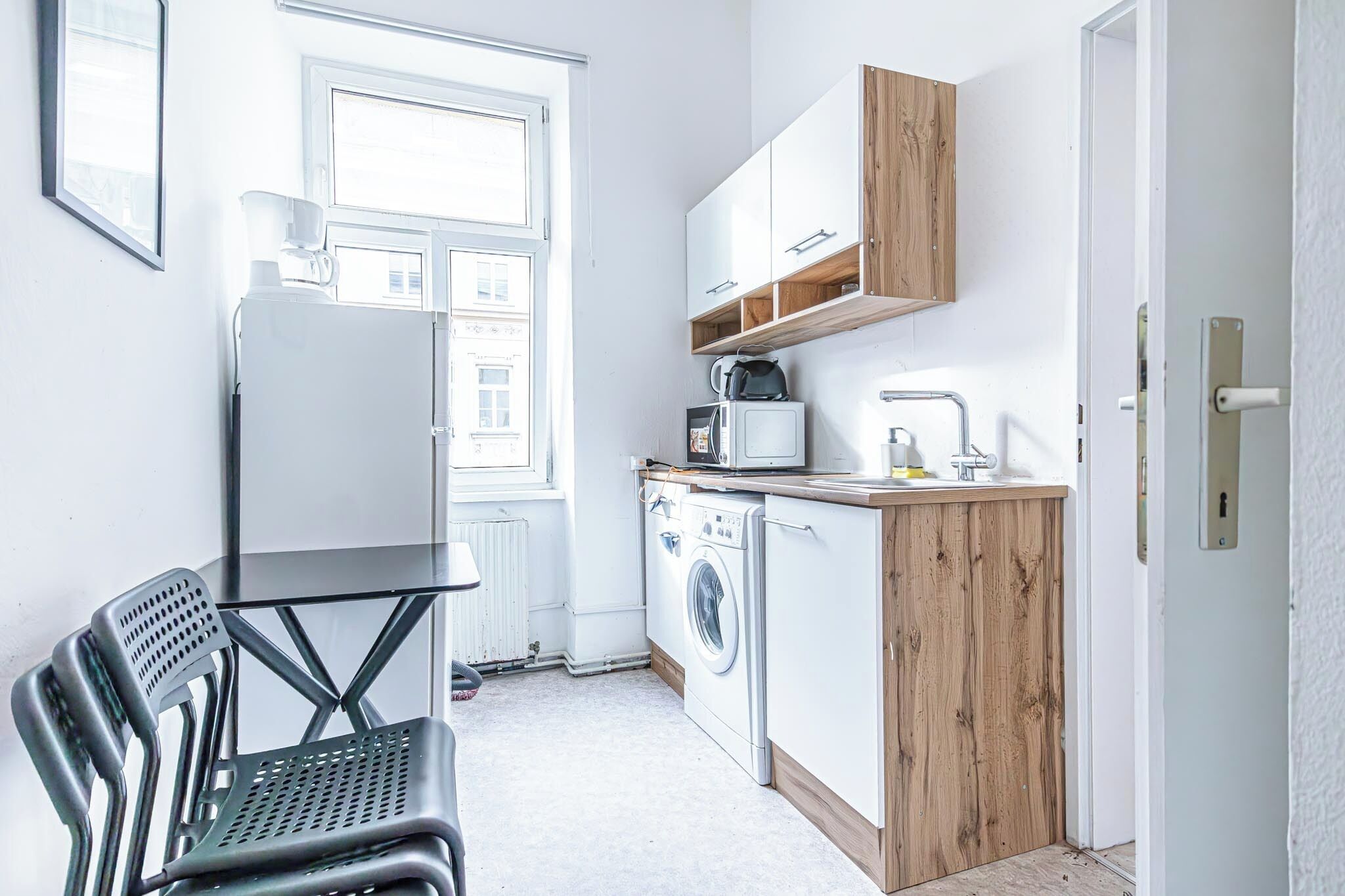 Een kleine keuken met een wasmachine, een magnetron en een koffiezetapparaat. Er is een raam dat natuurlijk licht biedt, en een deur die naar een andere kamer leidt.