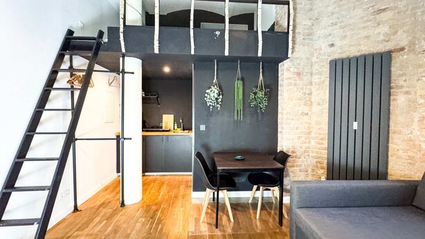 Un espacio habitable tipo loft con piso de madera, un área de comedor con mesa y sillas, un sofá gris, una escalera negra y una zona de cocina con planta colgante.