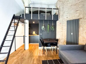 A loft-style living space with a wooden floor, a dining area with a table and chairs, a gray sofa, a black ladder, and a kitchen area with a hanging plant. - Cozy Studio for 4 on Prenzlauer Allee (Berlin)