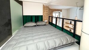 Una cama con colcha gris, dos almohadas, una pared de ladrillo y techo blanco.
