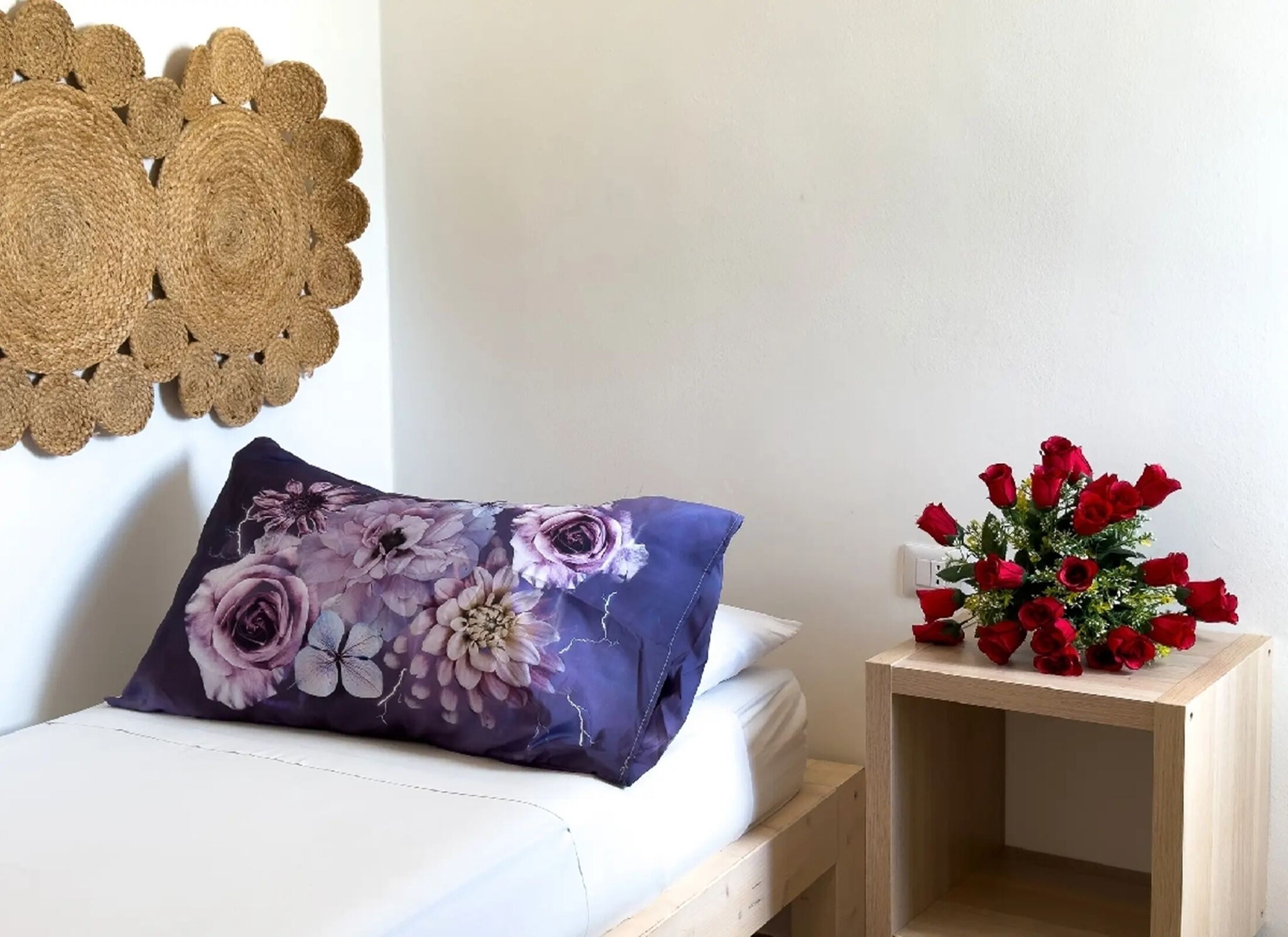 A bed with a floral pillow, a small wooden nightstand with a bouquet of red roses, and a decorative wall hanging.