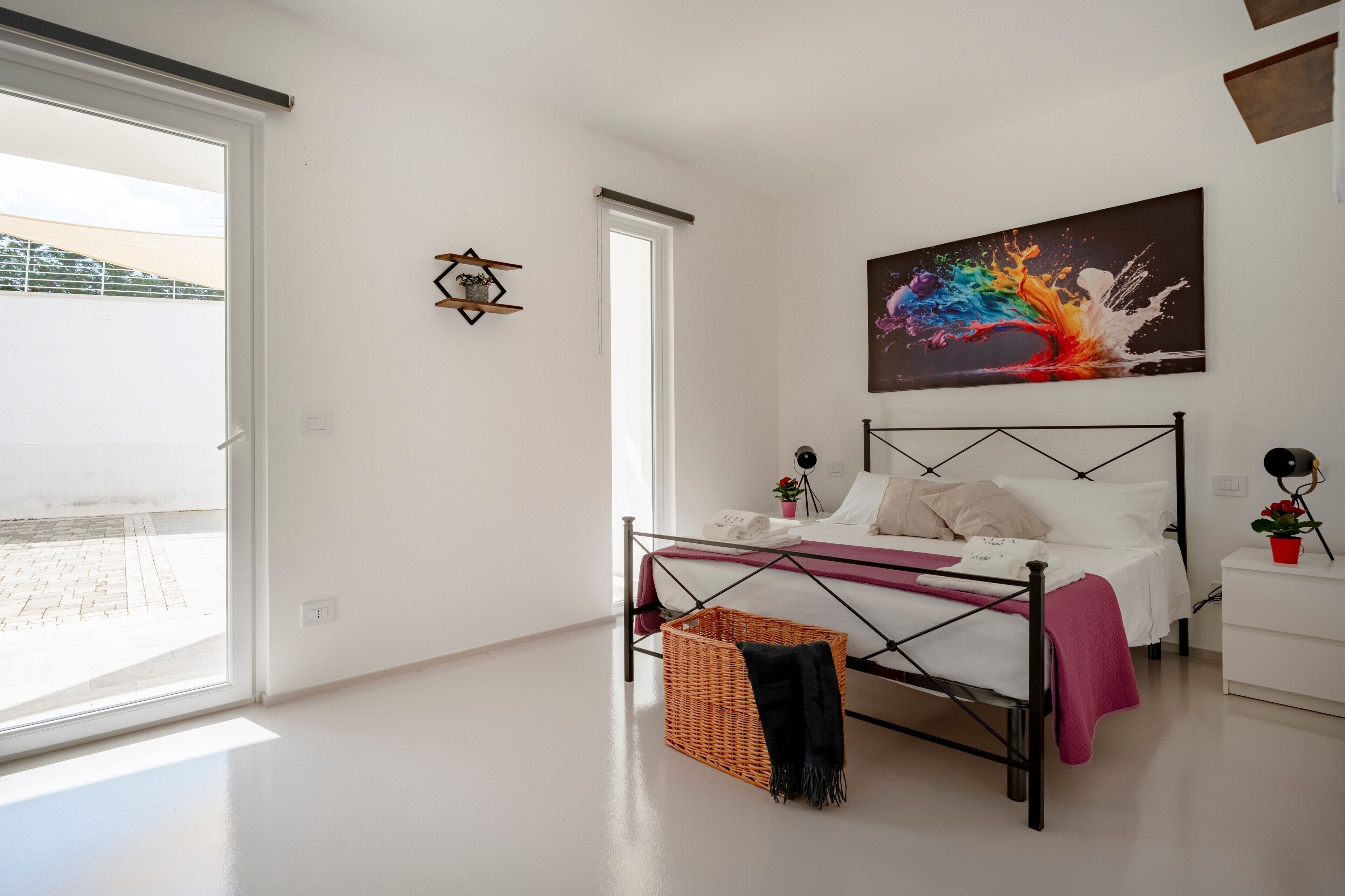 Een slaapkamer met een bed, een raam, een schilderij, een nachtkastje en een mand.