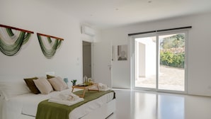 Ein modernes Schlafzimmer mit Bett, Nachttisch, grüner Tagesdecke und Blick durch eine Schiebetür auf einen Außenbereich.