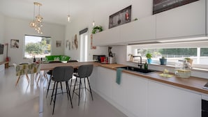 Eine moderne Küche mit einer zentralen Kochinsel, Barhocker, einem Essbereich mit einem grünen Sofa und einem großen Fenster mit Blick.