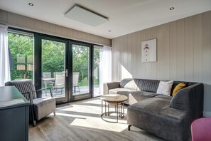 A modern living room with a grey sofa, a small round table, and a desk with a chair. Large glass doors lead to an outdoor patio with a table and chairs. - Spacious Holiday Home in Julianadorp (Julianadorp)