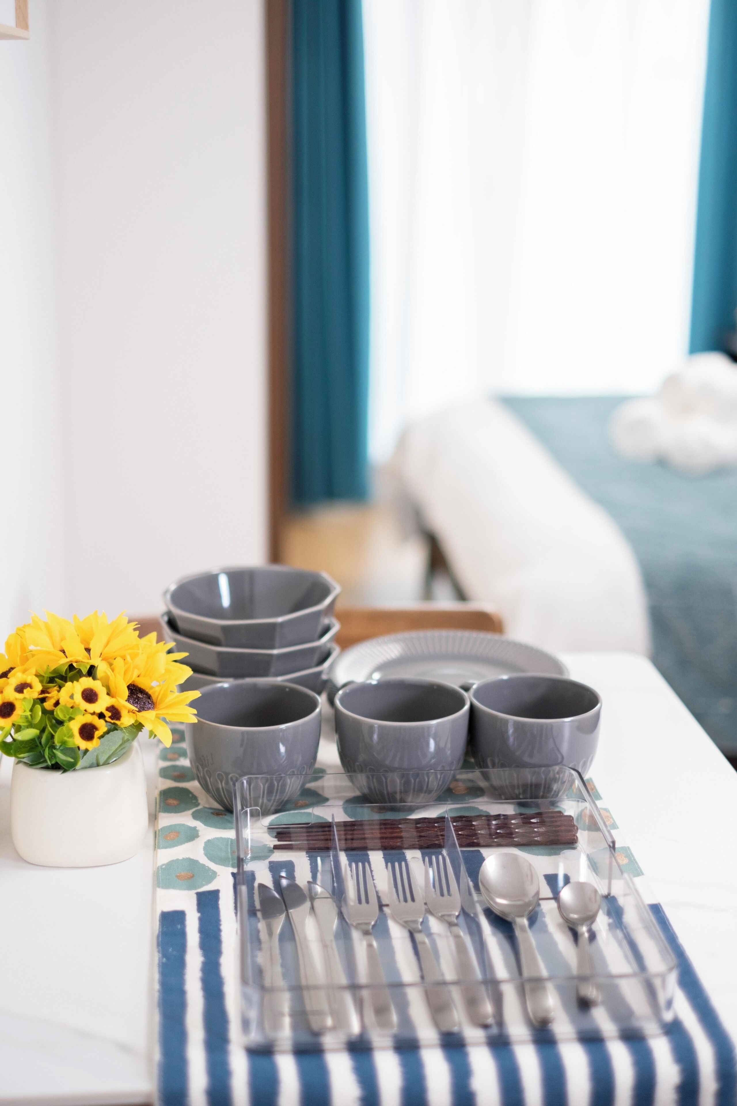 A neatly set dining table with grey bowls, spoons, forks, and a clear glass set.