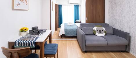 Une chambre d’hôtel moderne avec un plancher en bois, un canapé gris agrémenté d’un coussin à l’effigie d’un koala, une table à manger avec des chaises bleues et un lit recouvert d’une courtepointe bleue.