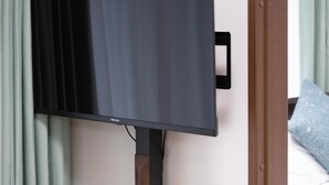 Une chambre d’hôtel moderne équipée d’un téléviseur à écran plat fixé au mur, d’une table de chevet en bois ornée d’un vase de fleurs et d’une assiette décorative, et d’un lit avec un oreiller bleu.