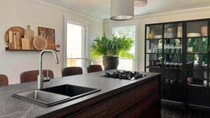 A modern kitchen with a large island, a built-in sink, wooden cutting boards, a potted plant, and a black cabinet with glassware. - Months & Seasons - Historic 1860s Estate - Daylesford (Daylesford - Hepburn Springs)