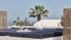 A rooftop lounge area with a view of the sea, featuring a cushioned seating area, a tray with a glass and a bottle, and wicker furniture.