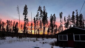 A cabin with a snow-covered ground, surrounded by tall trees and a sunset in the background.
