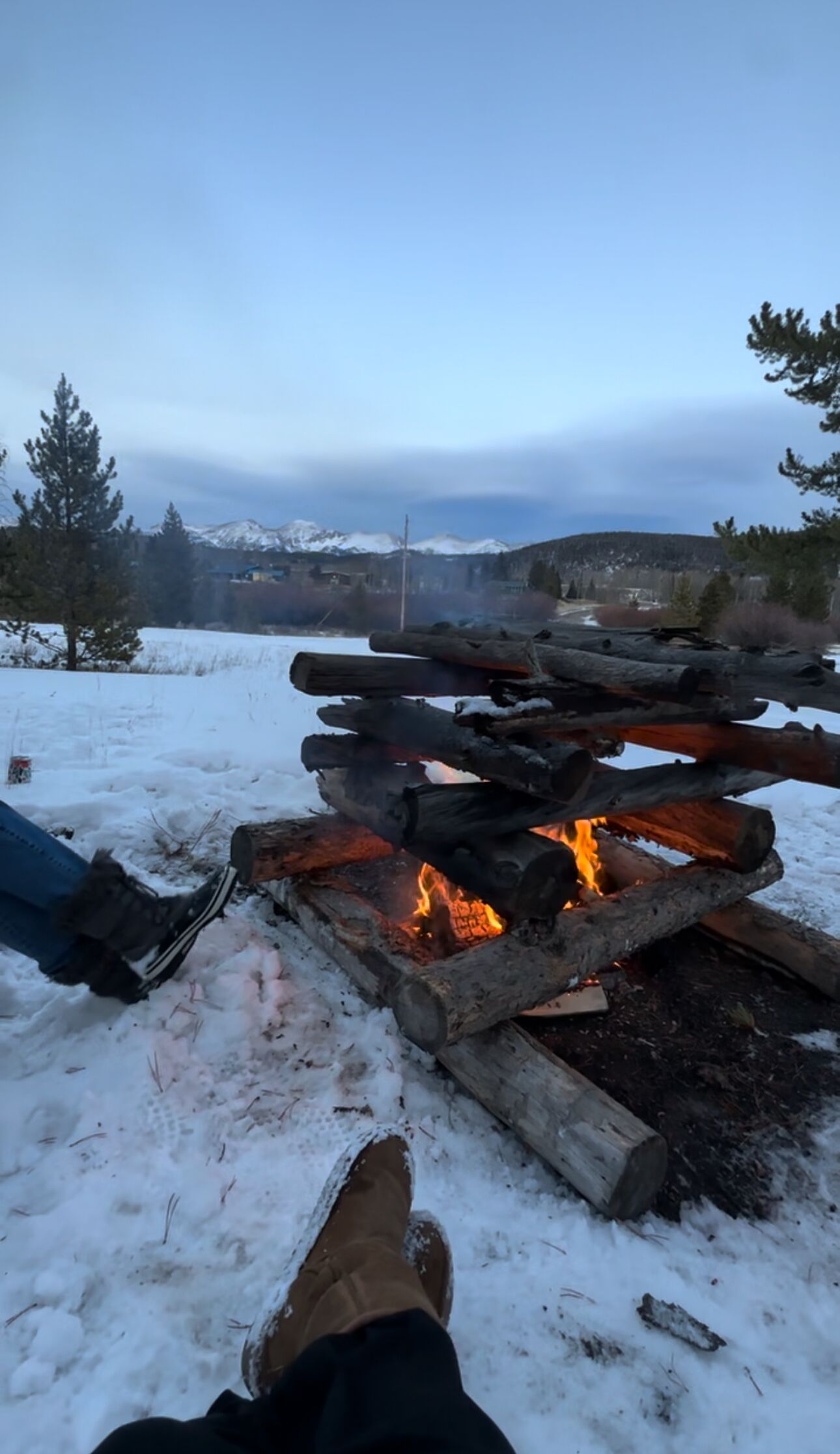 A person sitting by a campfire with snow-covered mountains in the background.