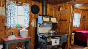 A cabin interior with a wood stove, a table with a checkered tablecloth, and a bed with a red cover.